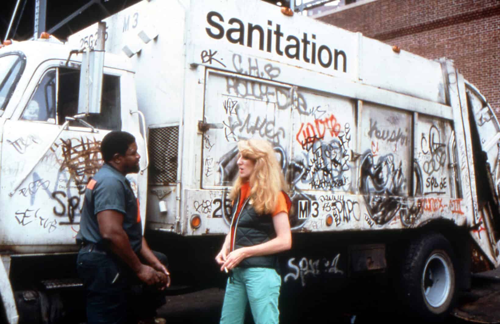 Graffiti-covered sanitation truck in urban setting with two people talking.