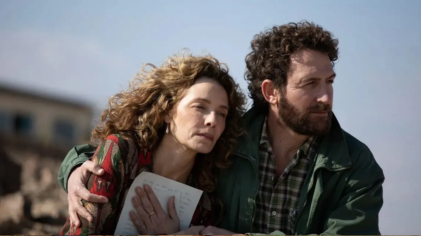 A medium shot of a man and a woman sitting close together outdoors under a clear, bright sky. The woman, with voluminous curly light-brown hair, leans against the man with a somber, pensive expression, clutching a handwritten letter to her chest. The man, who has dark curly hair and a beard, wears a green jacket over a plaid shirt and has his arm around her shoulder, looking off into the distance. The background is softly blurred, showing hints of a rugged, light-colored landscape.