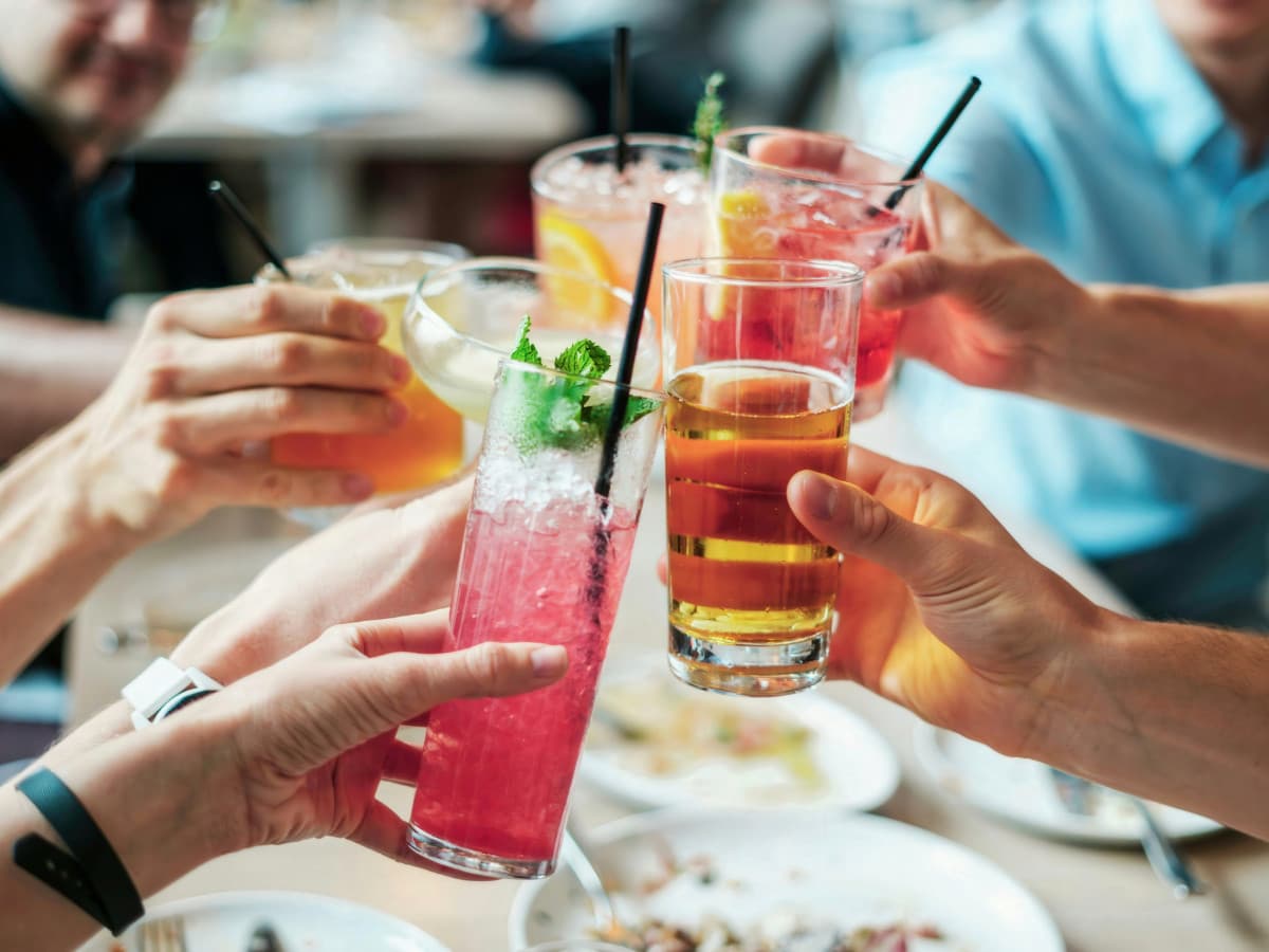 A close-up, eye-level shot capturing a group of people bringing their diverse drinks together in a celebratory toast above a restaurant table. In the foreground, two hands hold up distinctive glasses: one holds a tall, narrow glass filled with a vibrant pink cocktail, crushed ice, a black straw, and a fresh mint garnish; the other holds a clear glass of amber-colored beer or cider. In the background, other hands lift a variety of colorful cocktails, including an orange citrus drink, a yellow-tinted beverage, and a bright red cocktail with a lemon slice. The out-of-focus background shows glimpses of smiling faces, a person in a light blue shirt, and plates of food on the table, creating a warm, social atmosphere.