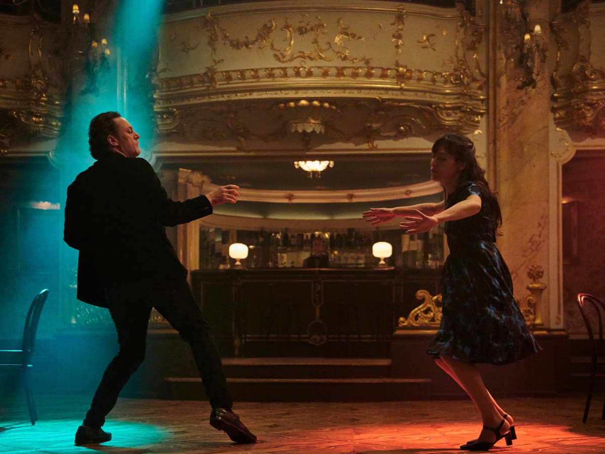 An athletic-looking man and a woman are captured mid-dance in the center of an ornate, dimly lit ballroom or theater. The man, on the left, is wearing a black suit and is leaned back on one leg with his arms outstretched, caught in a dynamic movement. The woman, on the right, wears a dark blue floral dress and is also in motion, her arms reaching toward him.