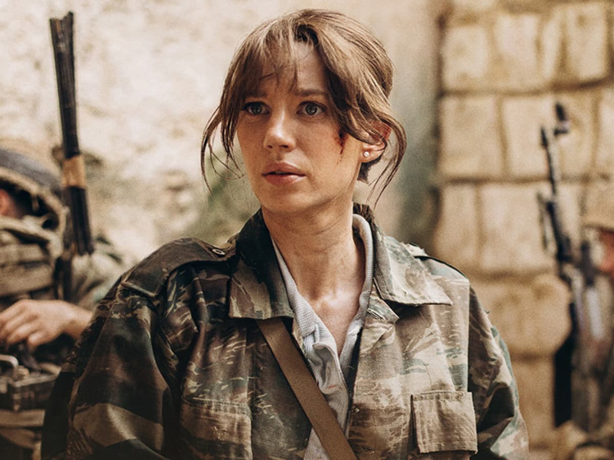 A close-up film still of a young woman with a concerned, intense expression. She has brown hair pulled back with loose strands framing her face, light blue eyes, and a small smudge of blood or dirt on her left cheek. She wears a camouflage military shirt open over a light grey top, with a brown canvas strap slung across her chest. She stands in a gritty, warm-toned outdoor setting with textured stone walls in the background. Part of another person in military gear and equipment is visible out of focus behind her on the left.