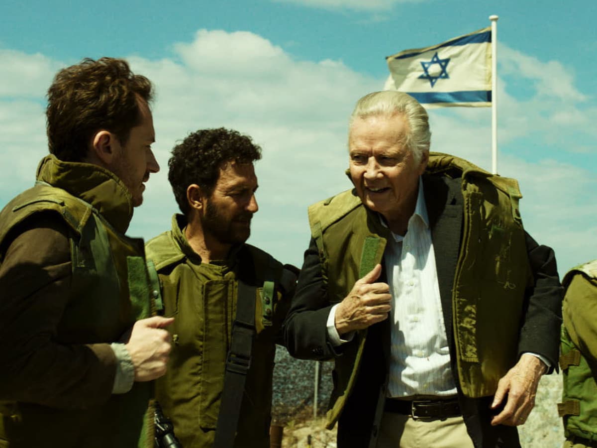 A wide film still showing an older, light-skinned man with swept-back white hair—resembling actor Jon Voight—smiling broadly as he is helped into a green military tactical vest. He wears a light, striped dress shirt, khaki trousers, and a dark blazer underneath the vest. He is surrounded by four younger men dressed in olive-green military uniforms. To his left, a man with dark curly hair and a beard assists him. To his right, another soldier with sunglasses watches on. In the background, an Israeli flag flies against a bright blue sky with scattered white clouds, and elements of a military outpost or checkpoint are visible. The overall color grading has a distinct warm, greenish-yellow tint.