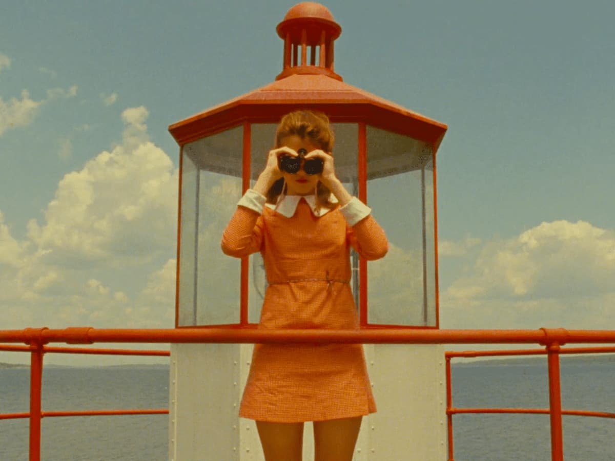 A centered, symmetrical medium shot of a young girl with reddish-brown hair standing on the balcony of a lighthouse. She wears a vintage pink-and-white checkered dress with a white Peter Pan collar and holds black binoculars to her eyes, looking directly toward the camera. She is framed perfectly within the glass-paned structure of the red-capped lighthouse lantern room. The color palette is warm and nostalgic, dominated by golden yellows, soft oranges, and a muted blue sky with fluffy white clouds in the background.