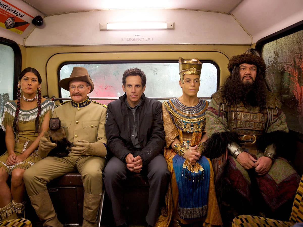 A movie still from Night at the Museum showing five characters and a small capuchin monkey sitting side-by-side on a bus seat. They are looking straight ahead with deadpan, serious expressions against a backdrop of a rainy, fogged-up bus window.