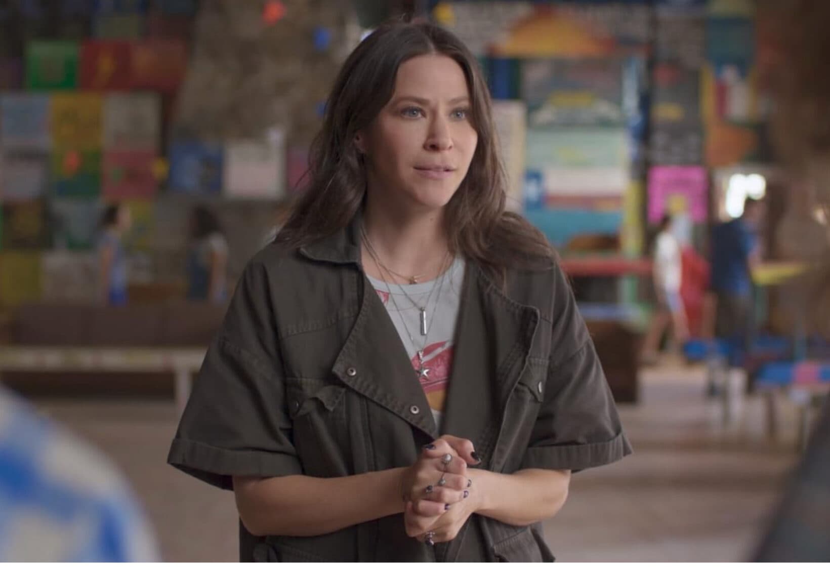 A medium shot of a woman with long brown hair, wearing a dark olive green short-sleeed utility jacket over a graphic t-shirt and layered necklaces. She has her hands clasped in front of her with a concerned or attentive expression. The background is a softly blurred indoor space featuring a colorful wall covered in various posters and signs, with a few out-of-focus figures walking in the distance.