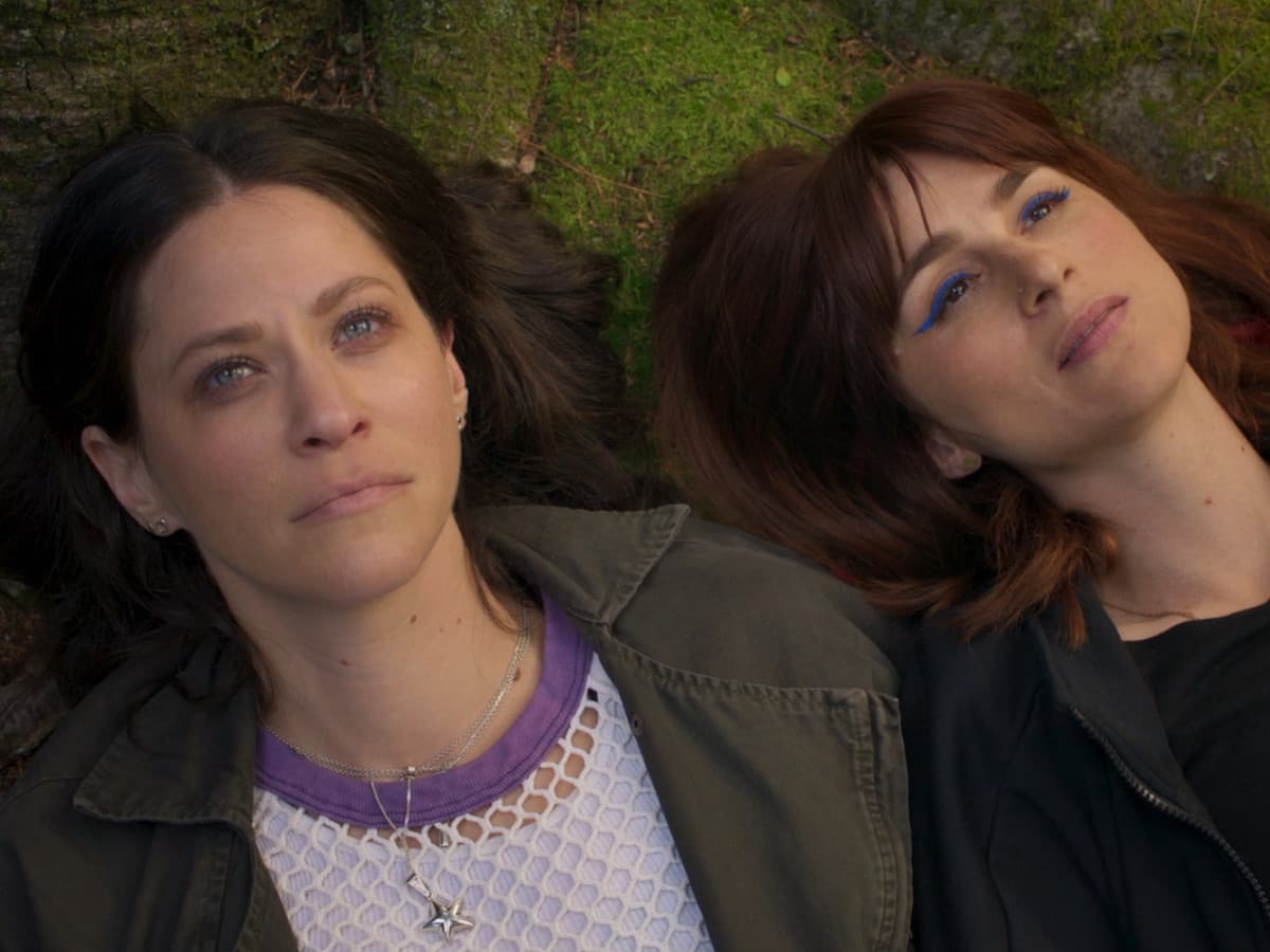 An overhead, close-up shot of two women lying down side-by-side on a patch of green moss and earth, their heads positioned close together. The woman on the left has dark brown, wavy hair framing her face. She has light-colored eyes, a reflective or somber expression, and a visible tear on her right cheek. She is wearing a purple-trimmed white mesh top under a dark olive green jacket, accessorized with a layered silver necklace featuring a star pendant. The woman on the right has auburn-brown hair with bangs. She is looking slightly upward and away from the camera with a serene expression. She wears bright, dramatic blue winged eyeliner and a black jacket. The lighting is soft and natural, emphasizing the texture of the mossy ground beneath them.