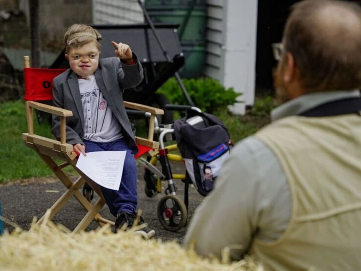 A young boy named Oliver, wearing a grey blazer over a graphic t-shirt, sits in a wooden director's chair outdoors. He holds a script in one hand and points forward confidently while directing. A yellow walker is visible behind his chair. In the foreground, an adult is seen from behind, listening to his direction.