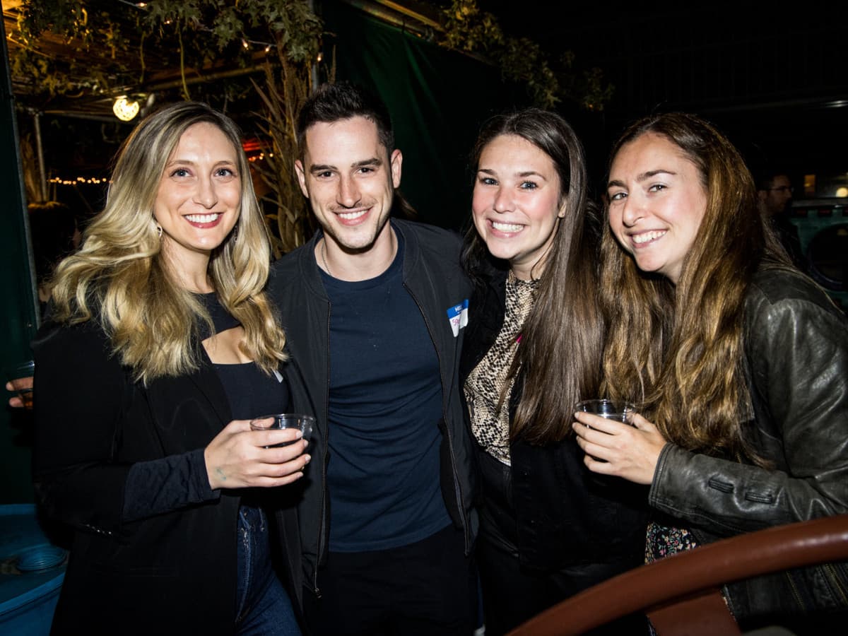Four young adults in their 20s and 30s smiling and holding drinks together at a night event inside a decorated sukkah.