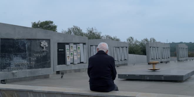 An elderly man, seen from behind, sits quietly before a large outdoor stone memorial featuring Hebrew inscriptions under an overcast gray sky.