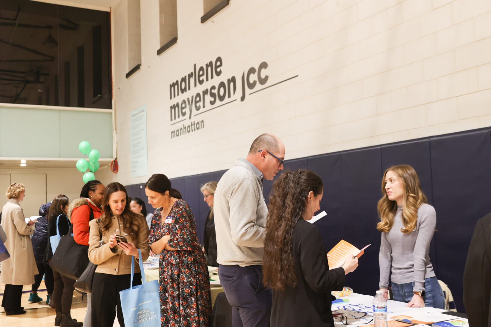 Attendees speak with exhibitors and browse resources at the Marlene Meyerson JCC Manhattan Special Education School Fair, with information tables and the JCC logo visible in the gym.