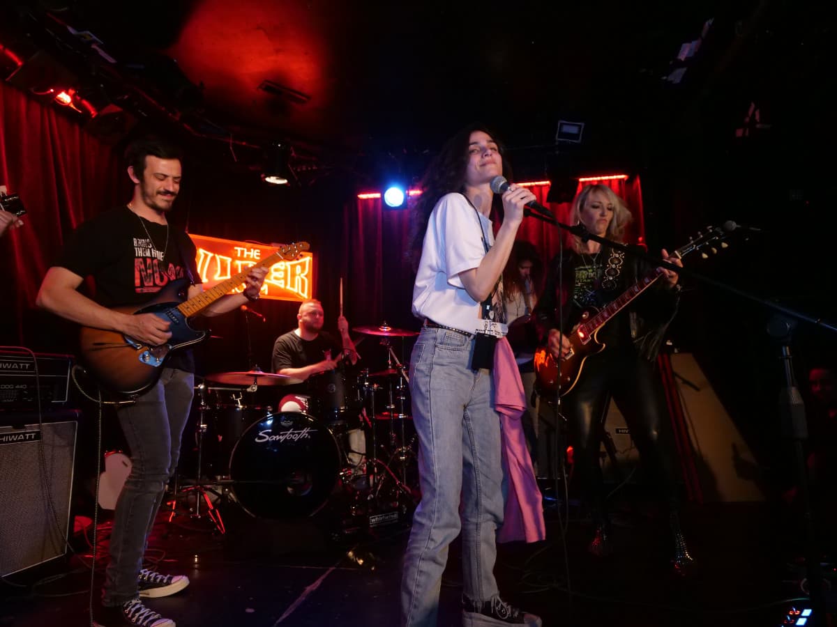 A live band performs on stage at a dimly lit venue under red and blue stage lights. In the foreground, a woman with long dark hair, wearing a white t-shirt and light-wash jeans, sings into a microphone. To the left, a man plays a sunburst electric guitar, while a drummer sits behind a black Sawtooth drum kit in the background. On the right, a blonde woman in a leather jacket plays an electric guitar. A red neon sign reading "The Viper Room" is visible on the wall behind the band.