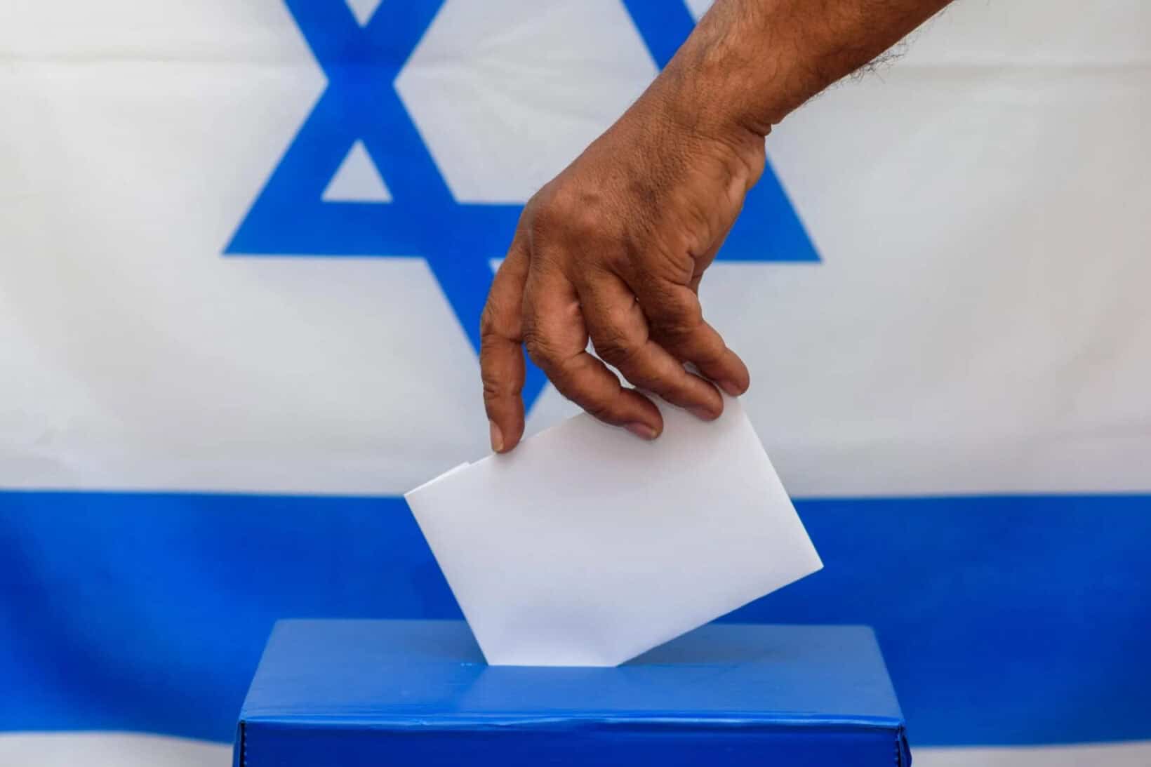 A hand places a ballot into a blue voting box in front of an Israeli flag with the Star of David.