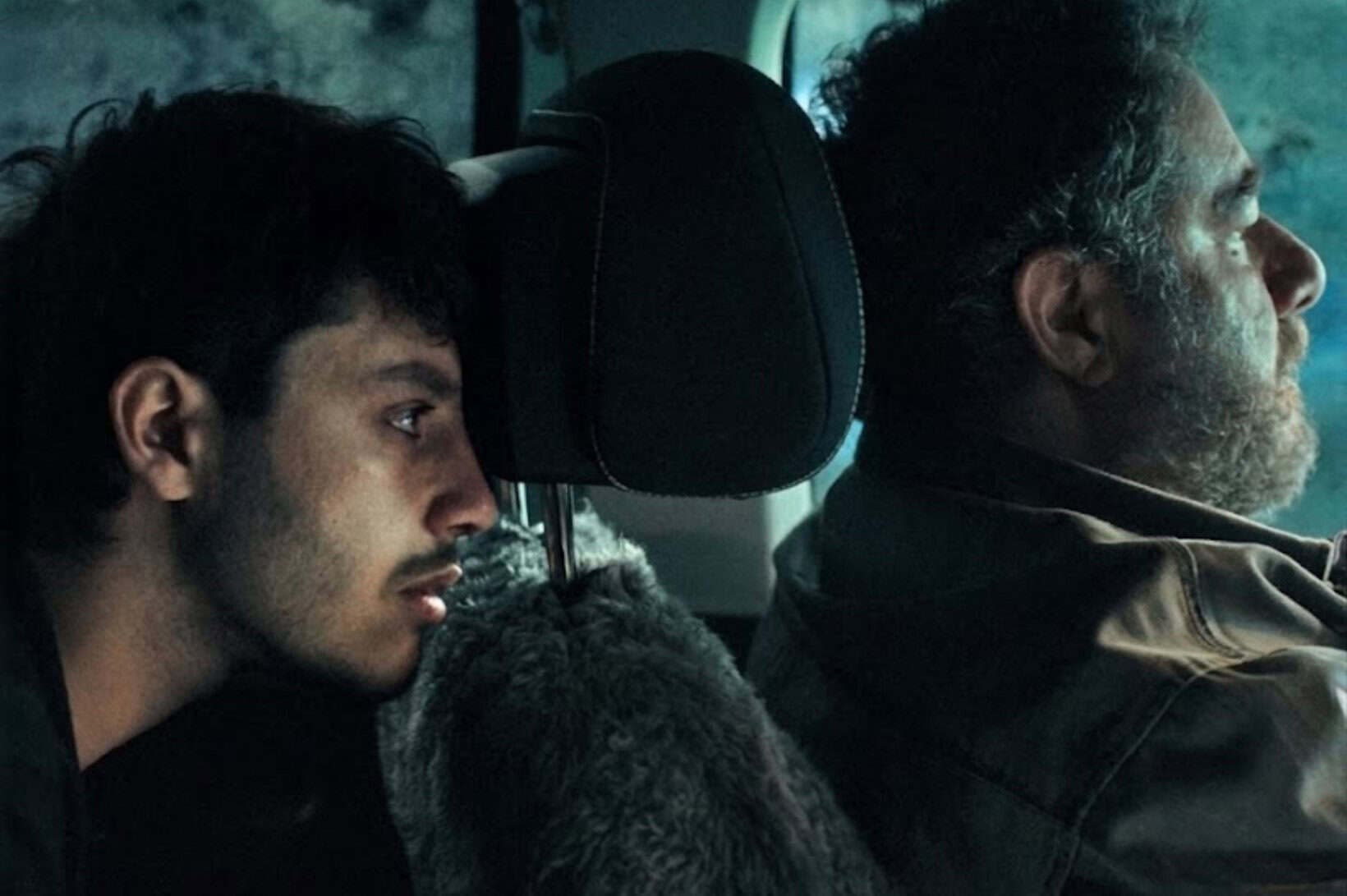 Cinematic still showing two men inside a car lit with cool blue evening light. On the left, a young man with dark hair and a light beard leans his face against the front seat headrest, looking solemn. On the right, an older man with a grey-ing beard sits in the front seat facing forward.