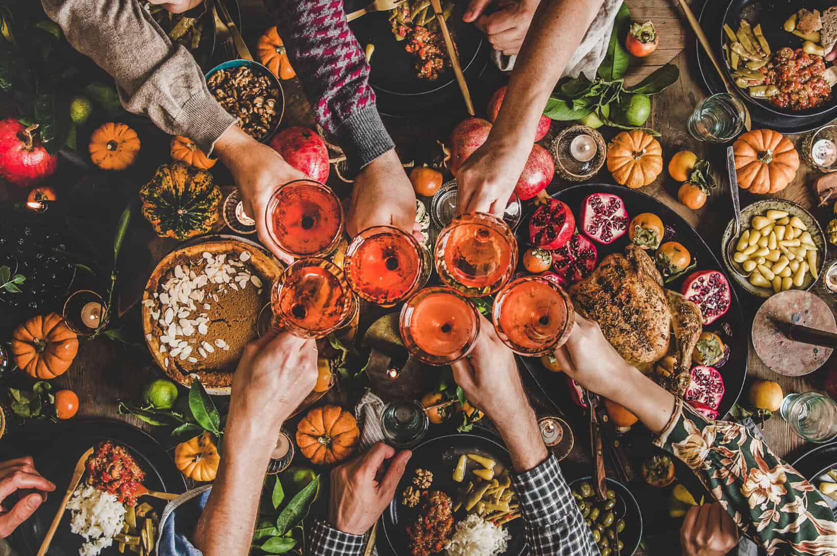 Overhead view of a festive Friendsgiving dinner table filled with autumn dishes, including a baked apple tart, roasted Brussels sprouts, soup bowls, cheese boards, lit candles, and small pumpkins, with several hands reaching in to share the meal.