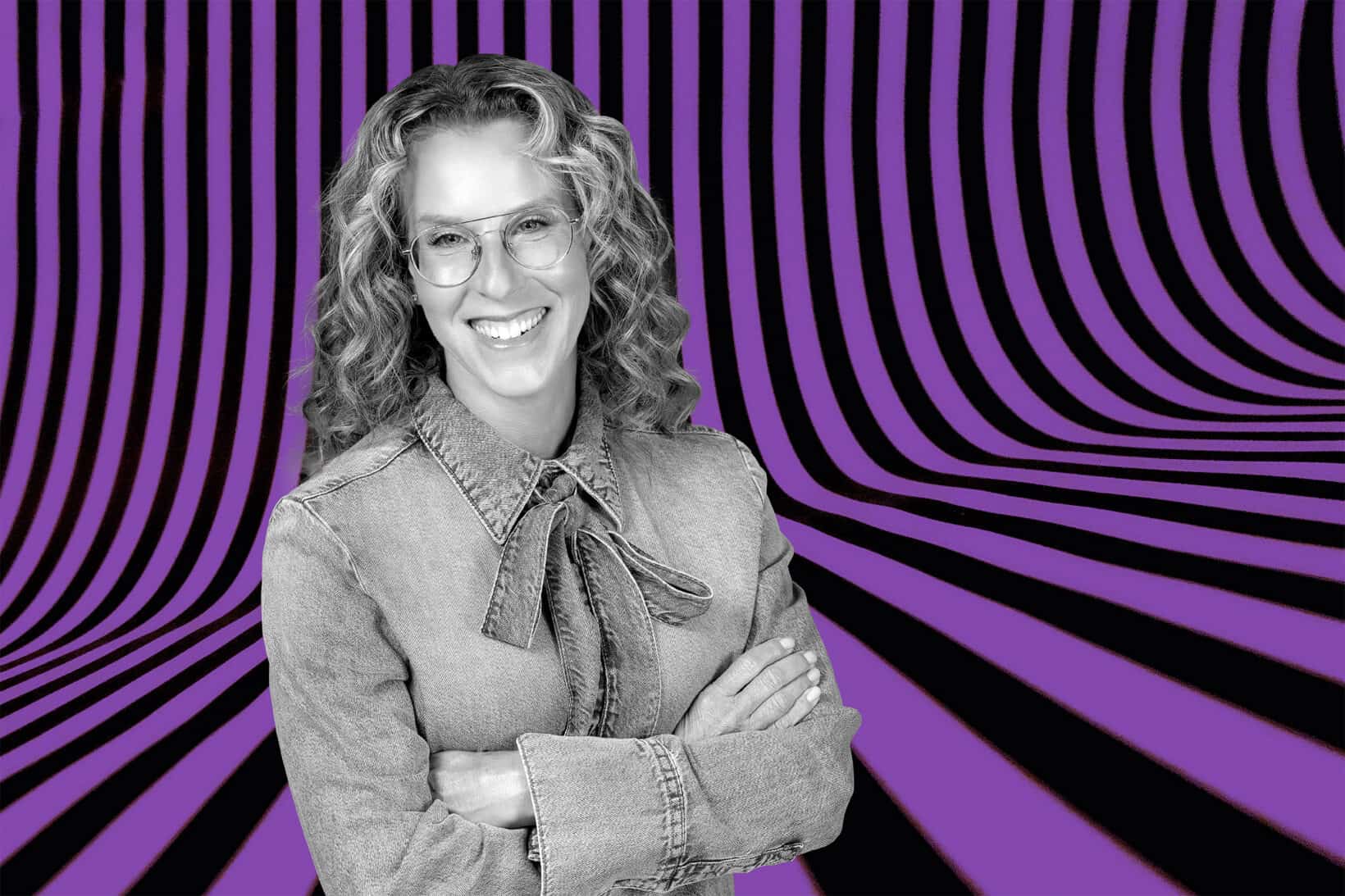 Black-and-white portrait of comedian Revital Vitelzon Jacobs with curly hair and glasses, standing with her arms crossed against a bold purple-and-black wavy patterned background.