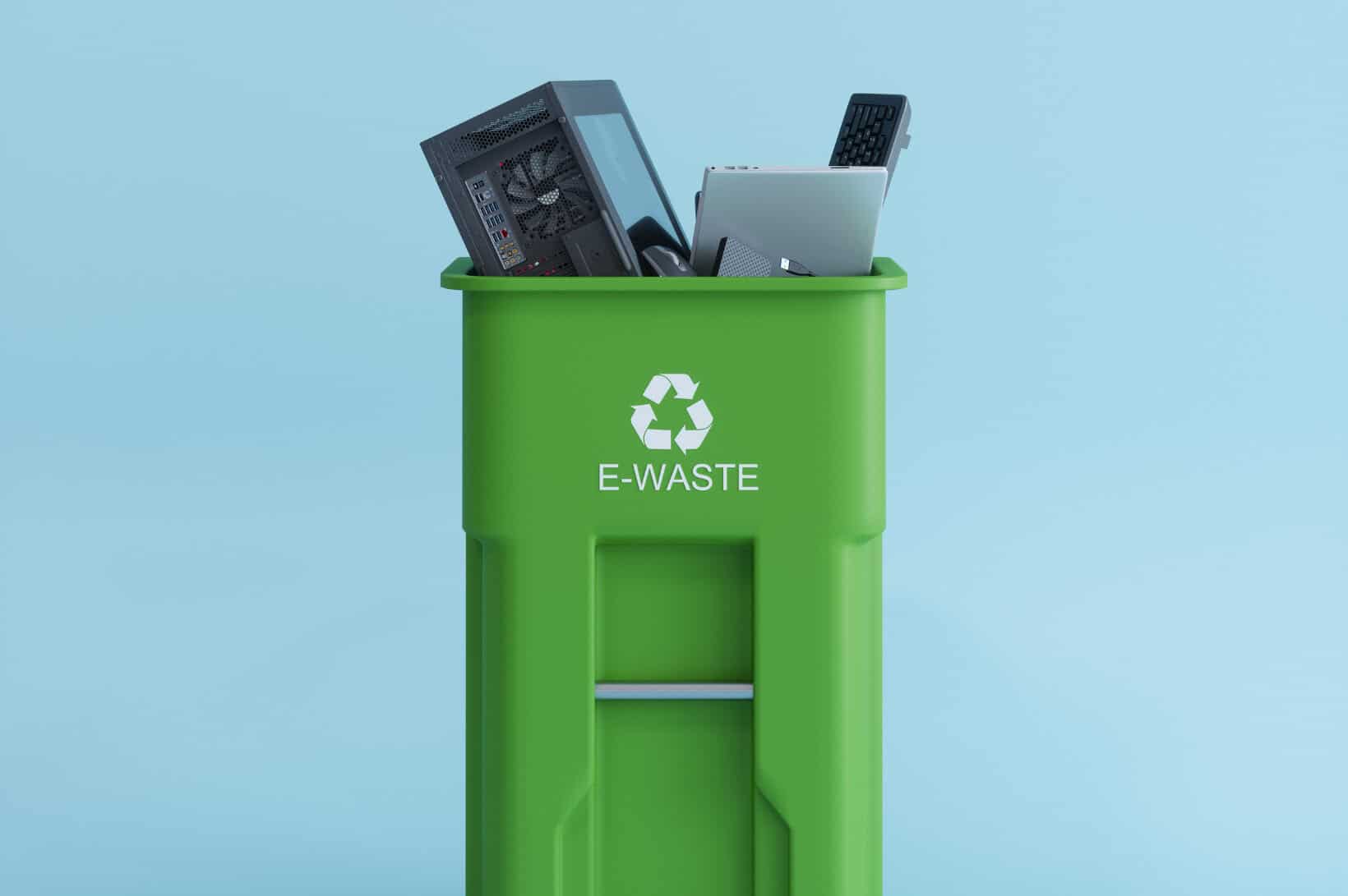 A tall green recycling bin labeled "E-WASTE" with a white recycling symbol, filled with discarded electronic devices including a computer tower, laptop, keyboard, and computer mouse against a solid light blue background.
