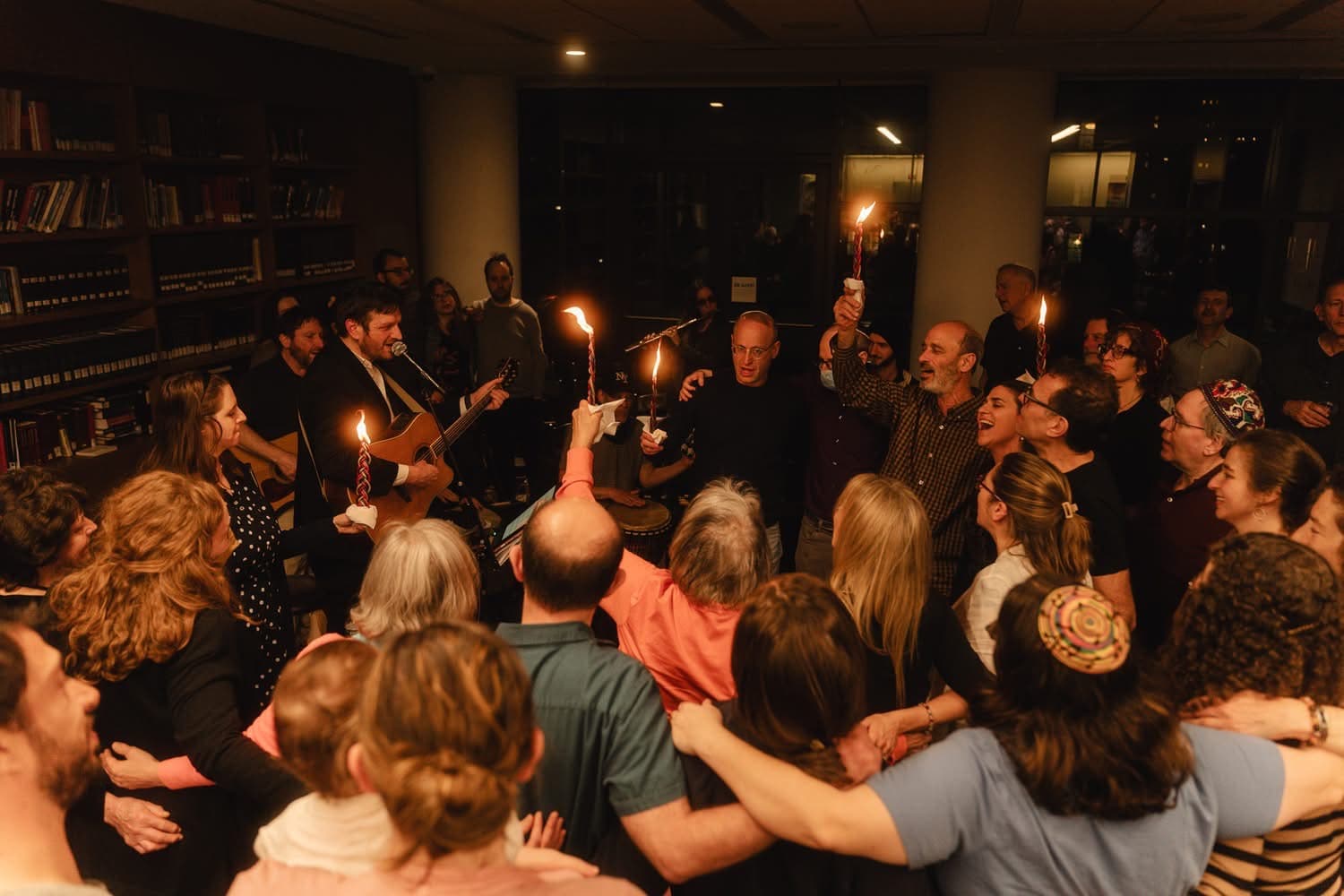 A warm, candlelit room filled with a large, diverse group of people standing in a close circle with their arms around each other. In the center, a musician plays an acoustic guitar and sings into a microphone, while others hold aloft multi-wicked Havdalah candles and a drum. The atmosphere is intimate and joyful, capturing a communal moment of song and connection as they transition from Shabbat into the new week.