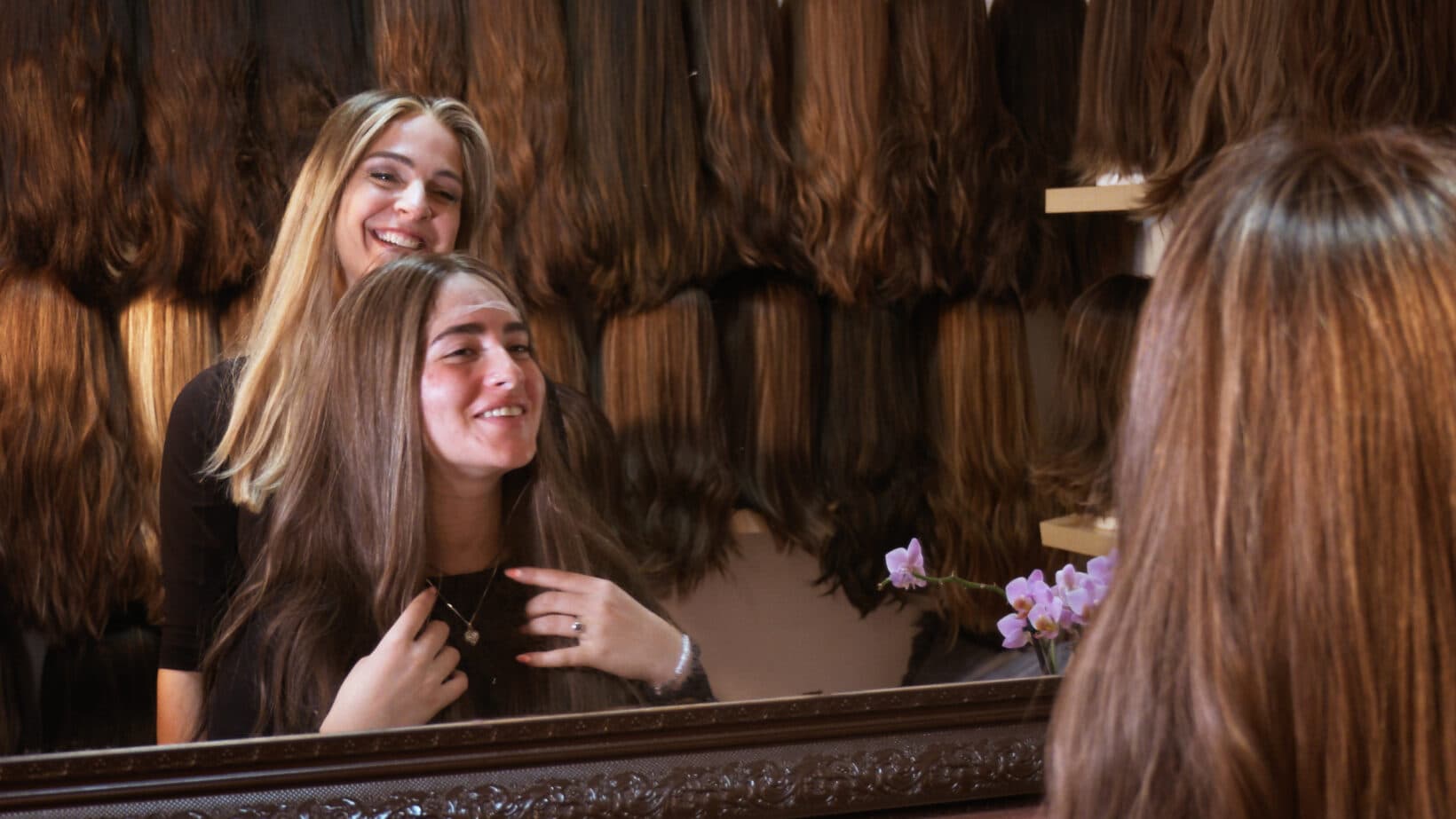 A medium shot of two women smiling in front of a mirror in a room filled with wigs. A woman with long blonde hair stands behind another woman with long brown hair, who is sitting and looking at her reflection in the mirror. Both women are smiling. In the background, numerous wigs in various shades of brown and blonde hang on the wall. In the bottom right corner, a purple orchid is visible in the reflection of the mirror.