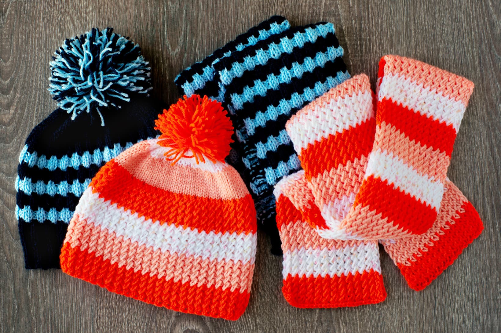 Flat lay view of two matching set knit beanie hats with pom-poms and scarves in striped navy/light blue and orange/white patterns, arranged on a dark wooden surface.