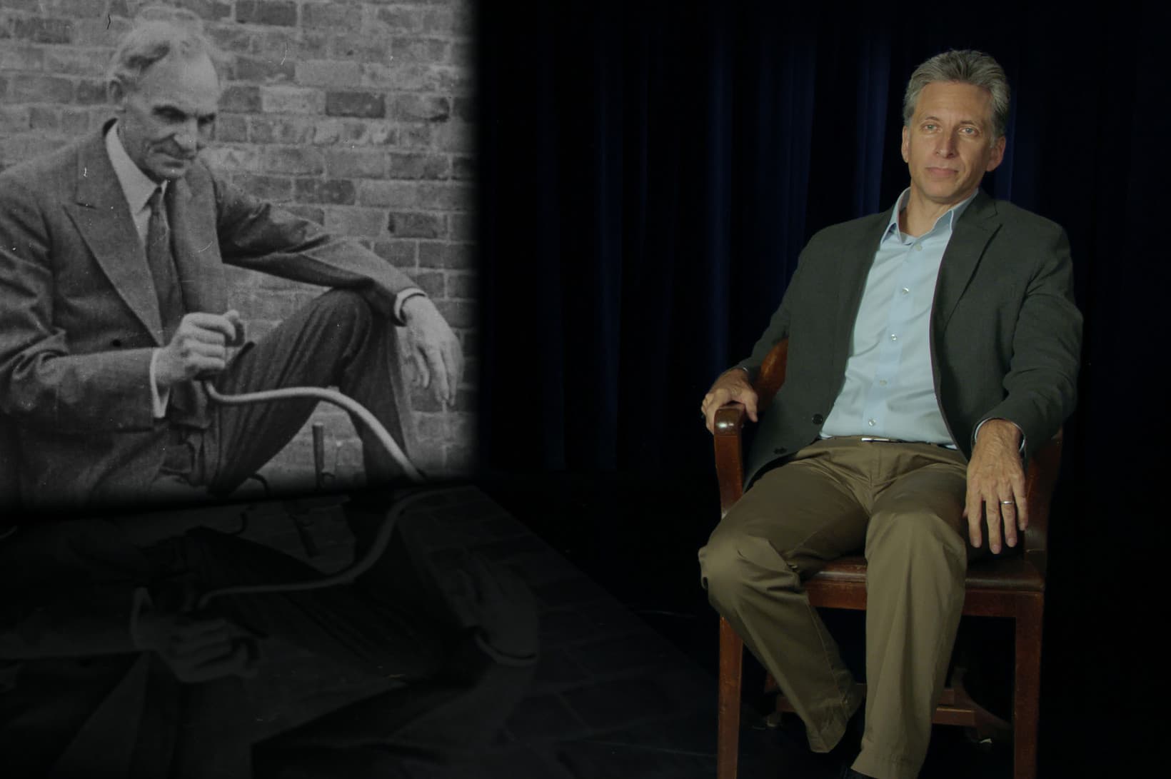 A man in a jacket sits in a studio on the right beside a vintage black-and-white photo of an older man crouching outdoors.